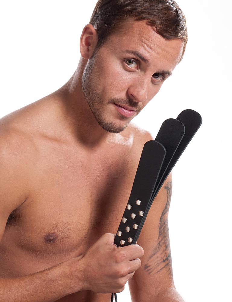 A muscular brunette man poses in front of a blank background, holding up the Kinklab Triple Leather Slapper.