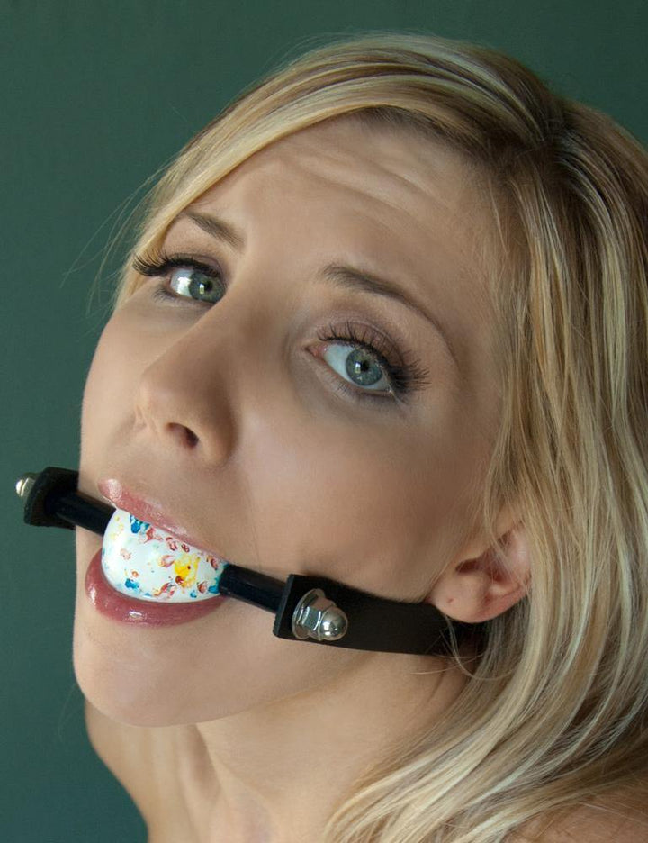  A close-up of a blonde woman wearing the Kinklab Jawbreaker Gag with a Black strap is shown. She looks upwards at the camera.