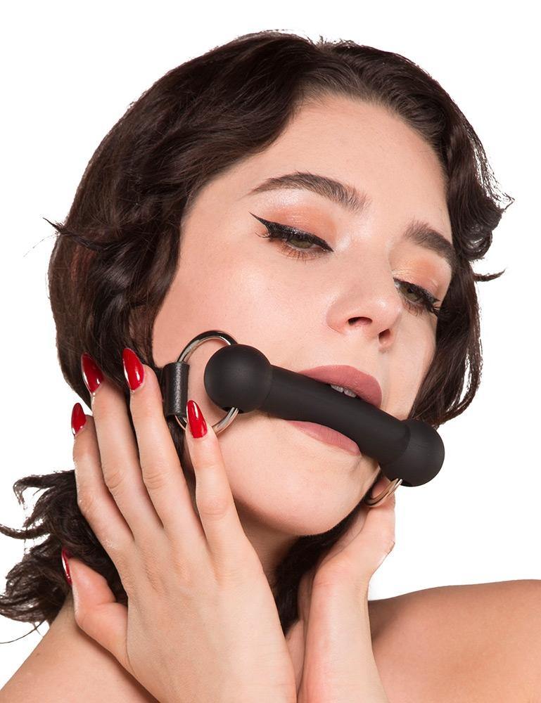 A woman with shoulder-length curly dark hair is shown against a blank background, wearing the Kinklab Silicone Bit Gag With A Leather Strap. Her nails are painted red, and her hands rest on her face below the gag. The gag is a rod made of black silicone.