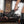 Load image into Gallery viewer, A woman&#39;s forearm and the side of her thigh in black latex stockings are shown in the foreground. In front of her is a man on a leather couch facing away from her. She holds the KinkLab Panamorphic Paddle 3-in-1 Spanking Set with the spikes on it.
