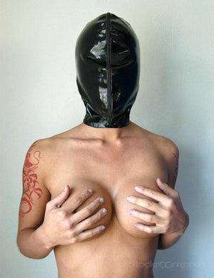 A topless woman is shown from the waist up, standing against a wall and covering her breasts with her hands. She wears the Extreme Rubber Hood zipped up, completely covering her face.