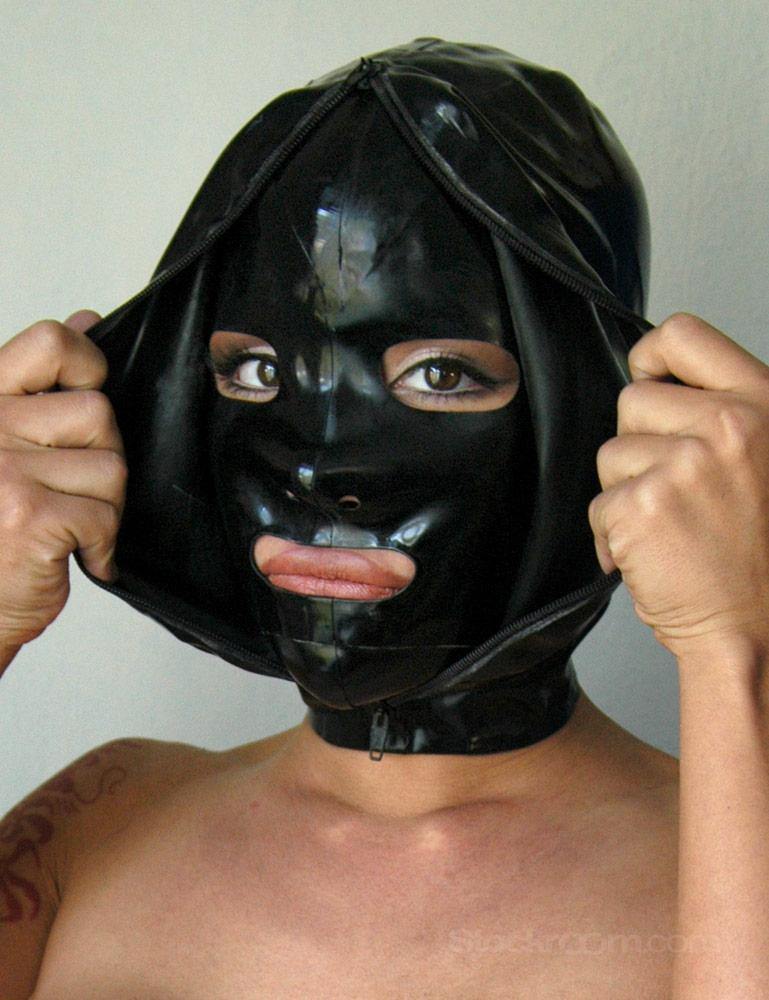 A close-up of a woman's face is shown wearing the black Extreme Rubber Hood. The hood is unzipped, and she pulls apart the outer layers to reveal the bank robber-style mask underneath. 