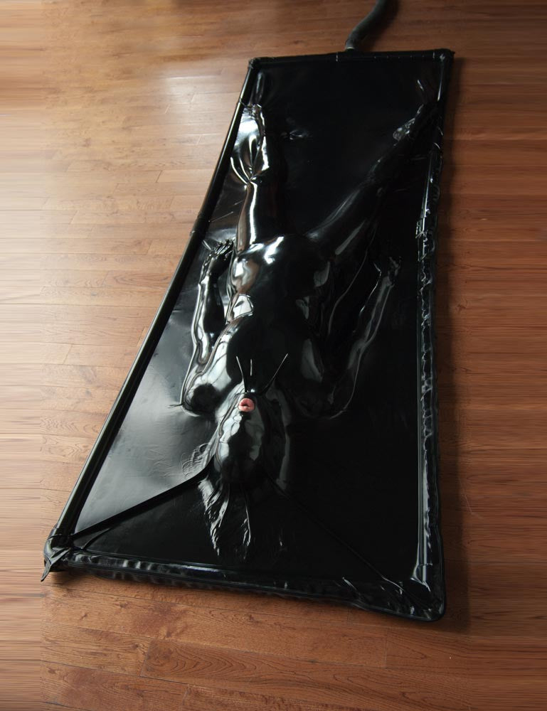  A woman is lying on her back in a Latex Vac-Bed on a wooden floor. The outline of the bed is black, and the latex is black. The woman is encased in the latex, which conforms to the exact shape of her body and seals her to the bed. Only her mouth is uncovered.