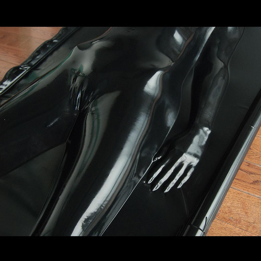 A close-up of the woman’s hips and thighs is shown in the black Latex Vac-Bed. The outline of her body is visible where the latex clings to it. 