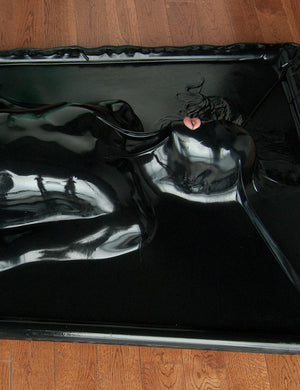  A close-up of the woman’s upper body in the black Latex Vac-Bed is shown. Her lips are pink and slightly parted. The latex is opaque and shows only the shape of her body. 