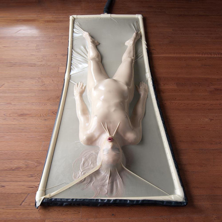 A woman is lying on her back in a Latex Vac-Bed on a wooden floor. The outline of the bed is black, and the latex is clear. The woman is encased in the latex, which conforms to the exact shape of her body and seals her to the bed. Only her mouth is uncovered. 