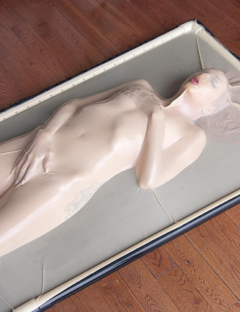 A nude woman is shown encased in the clear Latex Vac-Bed on a wooden floor. One of her hands rests on top of her vagina, and the other covers her breasts. The latex conforms to her body, which is mostly visible under the translucent latex.