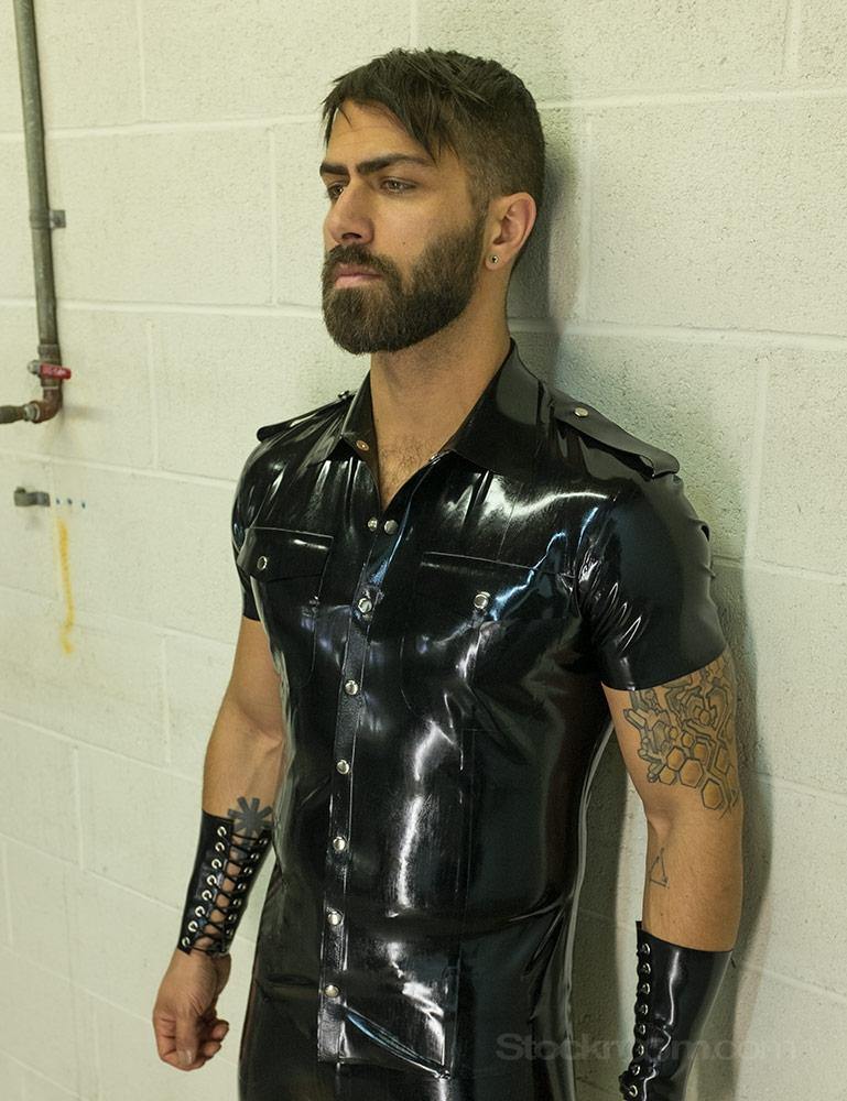 A brunette man with facial hair poses in front of a white wall. He wears the Uniform Shirt from Syren Latex in black, matching pants, and arm gauntlets. The shirt has metal snaps down the front, pockets on the chest, and a collar.