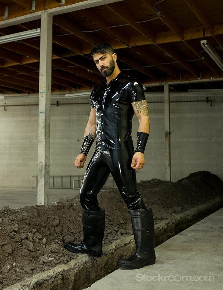 A brunette man with facial hair poses outdoors. He wears the Uniform Shirt from Syren Latex in black, matching pants and arm gauntlets, and large rubber boots.
