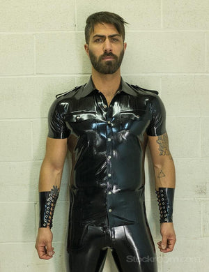 A brunette man with facial hair poses in front of a white wall. He wears the Uniform Shirt from Syren Latex in black, matching pants, and arm gauntlets. The shirt has metal snaps down the front, pockets on the chest, and a collar.