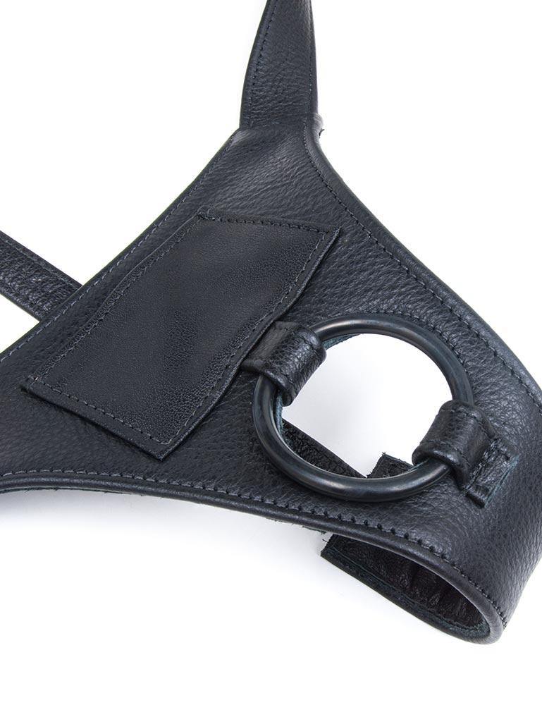 A close-up of the front piece of the black leather Buzz Me Tender strap-on harness is shown against a blank background. The harness has a black silicone O-ring. Above the harness is a small rectangular pocket for a vibrator.