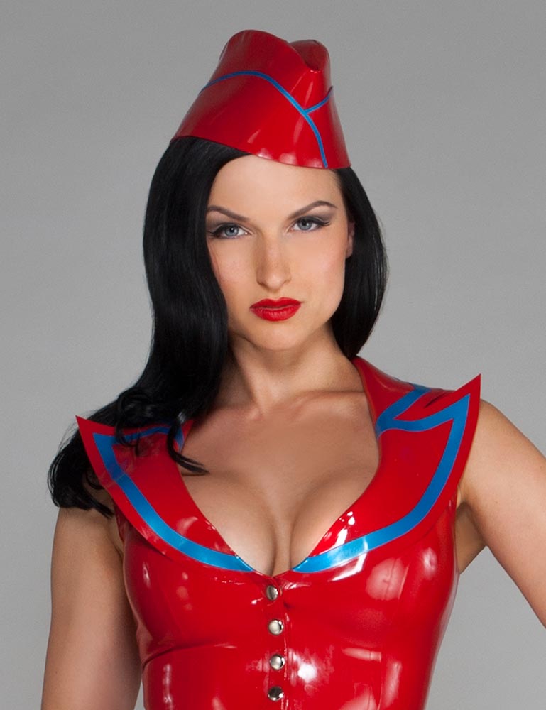 A woman with black hair and red lipstick wearing a red latex top poses in front of a grey background. She also wears the  Latex Garrison Cap by Syren latex in red with blue trim.