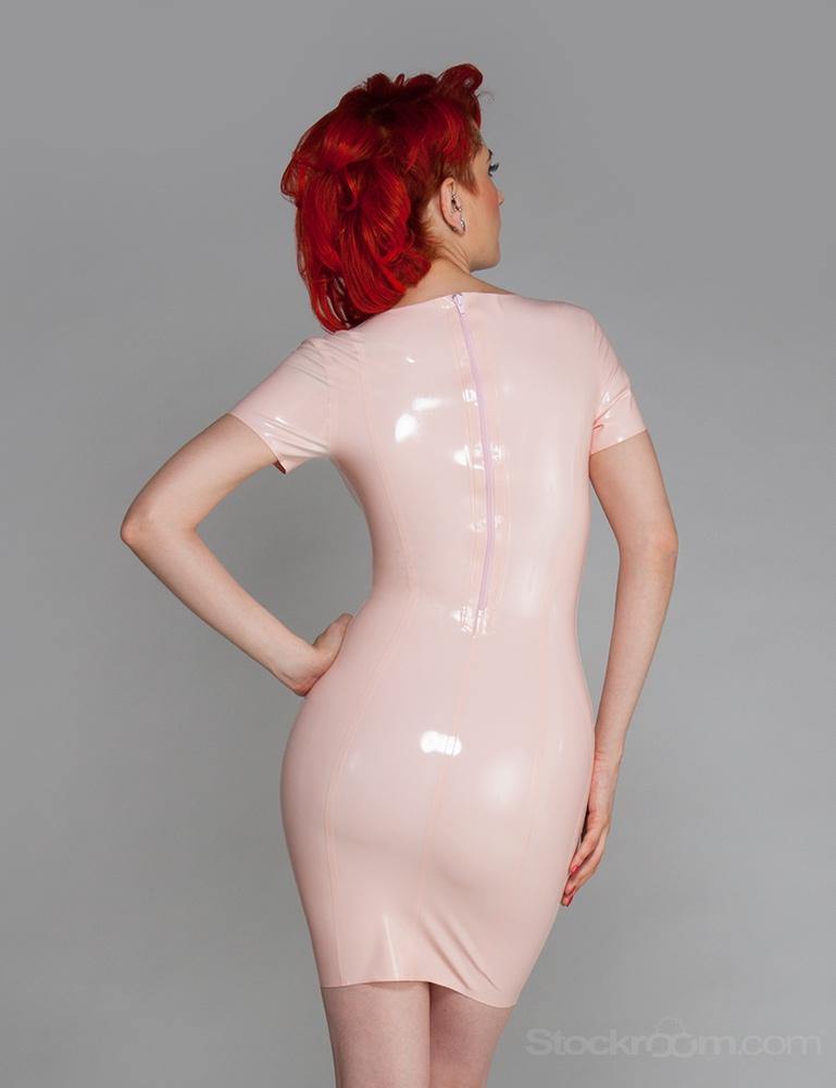 A woman with bright red hair poses in front of a grey backdrop, facing away from the camera. She wears the Latex Scoop Dress from Syren Latex in baby pink. The dress has a zipper down the back.