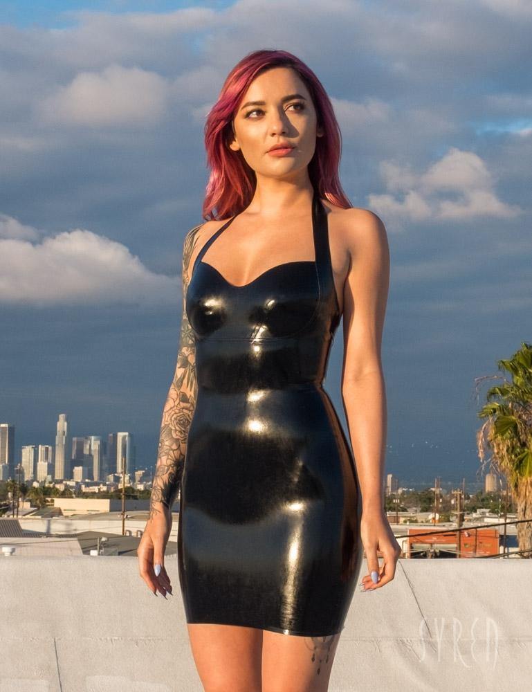 A woman with pink hair poses on a rooftop with a cloudy sky and the Los Angeles skyline behind her. She wears the Latex Halter Dress from Syren Latex in black.