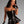 Load image into Gallery viewer, A brunette woman poses in front of a grey backdrop wearing the Rubber Laced Back Corset Dress with Ruffle Trim in black by Syren Latex. The strapless dress has a fitted corset top and a very short flared skirt at the hips.
