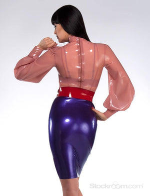 A woman with dark hair and red lipstick wearing a pink latex blouse and purple latex skirt poses in front of a grey backdrop, facing away from the camera. She wears a red latex Waist Cuff by Syren Latex.