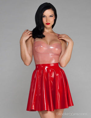 A dark-haired woman is posed in front of a grey backdrop. She wears a translucent pink latex camisole and a red latex Cheerleader Skirt from Syren latex.