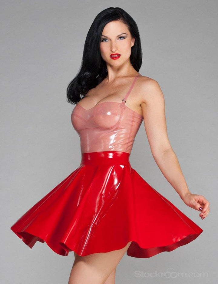 A dark-haired woman is posed in front of a grey backdrop. She wears a translucent pink latex camisole and a red latex Cheerleader Skirt from Syren latex.