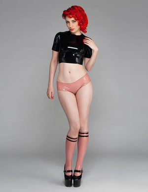 A woman with bright red hair poses in front of a grey backdrop. She wears a black latex crop top, pink latex panties, and the Varsity Socks by Syren Latex in transparent pink with black accents.
