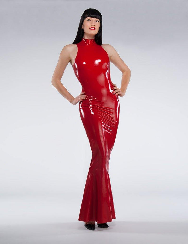 A brunette woman poses in front of a grey background. She wears the red fishtail latex gown from Syren Latex, which has a halter-style neck.