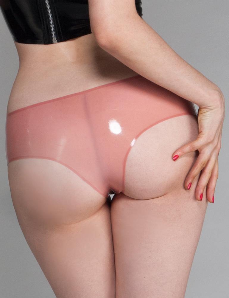 A close-up of a woman's butt and thighs is shown in front of a grey backdrop. She wears the Cheeky Panty by Syren Latex in rose pink. The panties are low-rise and expose some of her butt.