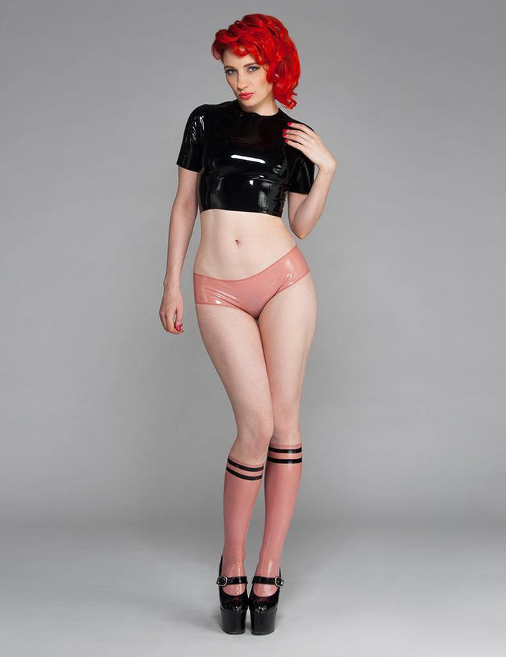 A woman with bright red hair poses in front of a grey backdrop wearing the Cheeky Panty by Syren Latex in rose pink. She also wears the latex zip-back crop top in black, athletic-style latex knee socks and black high heels.
