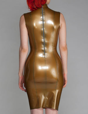 A woman with bright red hair poses in front of a grey backdrop, facing the wall, wearing the Tami Latex Dress by Syren Latex in Pearl Gold. The dress has a zipper down the back.
