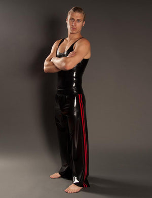 A blonde man poses in front of a grey background wearing the Men's Latex Track Pants by Syren Latex. The track pants are black with two red stripes on the sides. He also wears a black latex tank top.