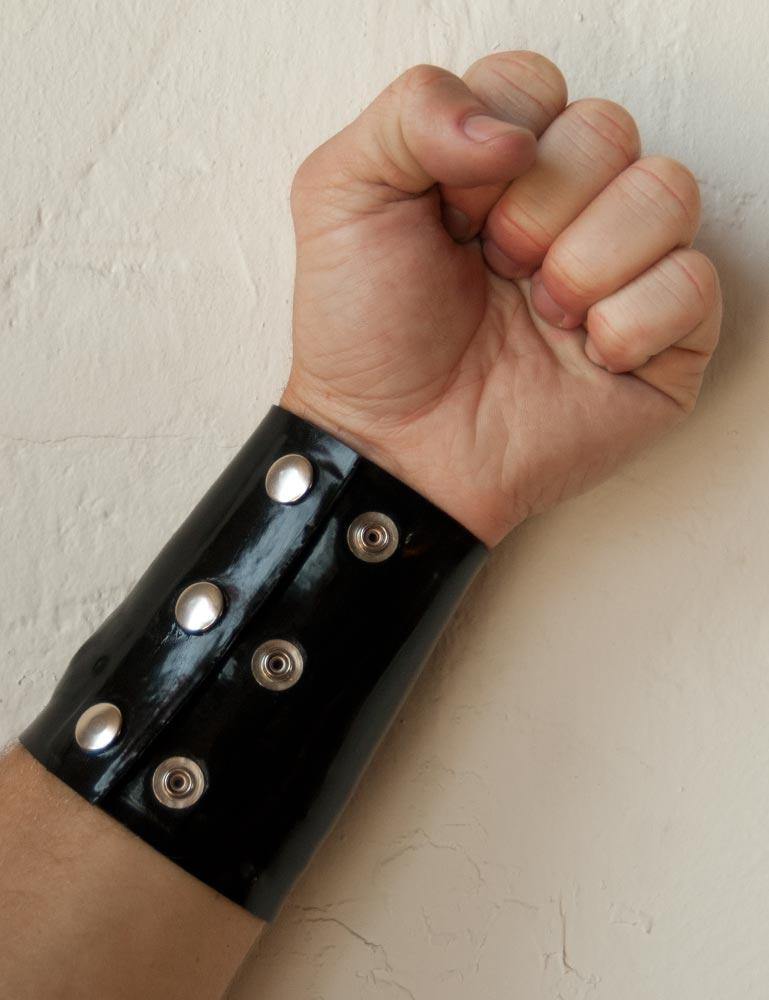 A close-up of a man's forearm is shown in front of a white wall. He wears the Latex Wrist Wallet by Syren Latex in black. The wallet resembles an armband and has two rows of three metal snaps.