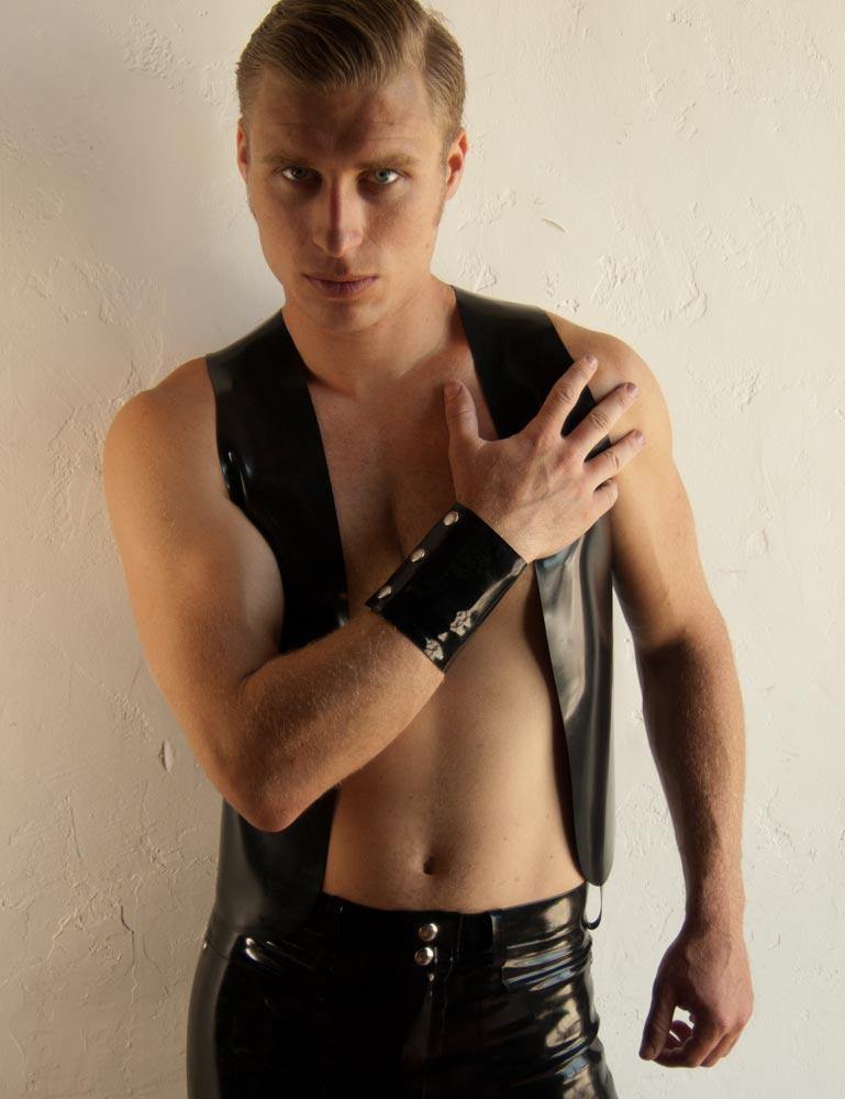 A blonde man wearing a black latex vest and no shirt poses in front of a white wall. He wears the Latex Wrist Wallet by Syren Latex in black. The wallet resembles an armband and has three metal snaps.