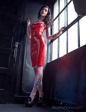 A photo of a brunette woman standing in front of an industrial window taken from an upwards angle. She wears the Newmar Latex Dress in red by Syren latex and black heels, as well as a clear PVC waist cincher. 