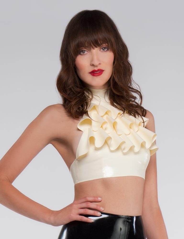 A brunette woman with red lipstick wearing the Jabot Halter Top by Syren Latex poses in front of a grey backdrop. The top is cream and is cropped to an inch under her bust. There are three rows of ruffles.