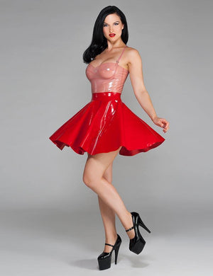 A woman with black hair and red lipstick wearing the Newmar Camisole by Syren Latex in transparent pink poses in front of a grey backdrop. She also wears a red latex cheerleader skirt and black high heels.