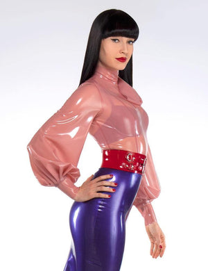 A woman with black hair and red lipstick poses in front of a grey backdrop. She wears the Garbo Blouse by Syren Latex in translucent pink, a purple latex pencil skirt, and a red latex waist cincher. Her bra is visible underneath her blouse.