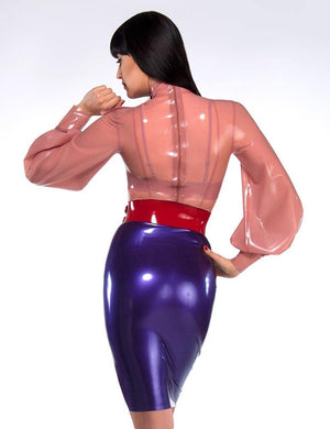 A woman with black hair poses in front of a grey backdrop, facing away from the camera. She wears the Garbo Blouse by Syren Latex in translucent pink, a purple latex pencil skirt, and a red latex waist cincher. The blouse has a zipper in the back.