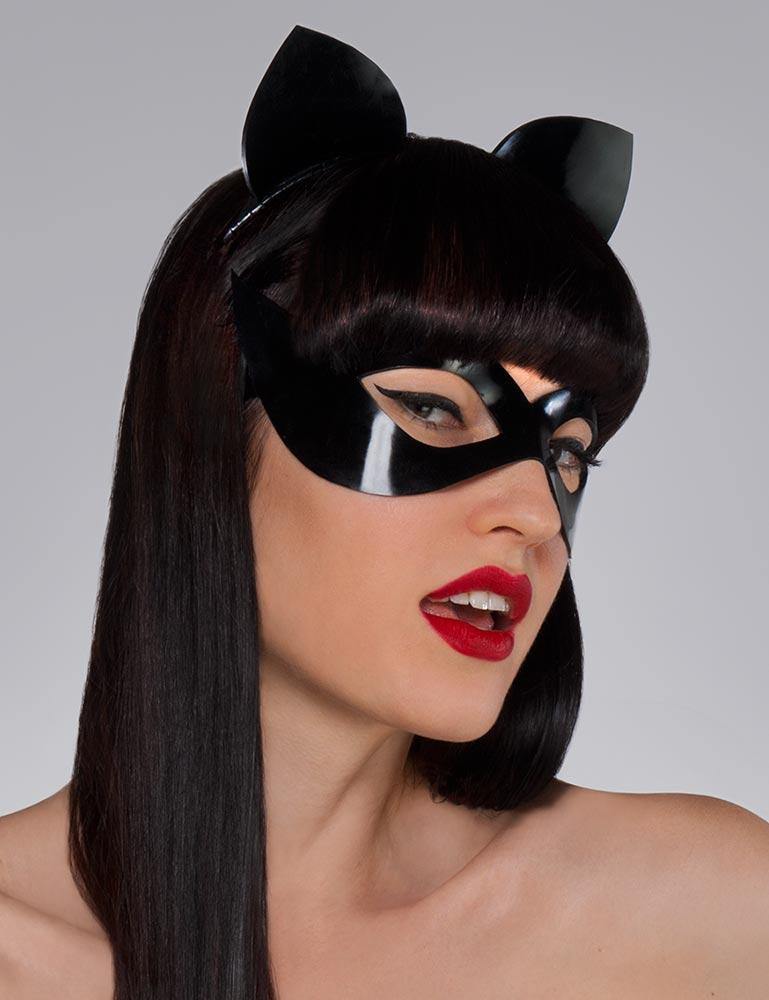 A headshot of a woman with dark hair and red lipstick posed in front of a grey backdrop. She wears the black Mistress Mask by Syren Latex, which outlines her eyes in a cat-eye style. She also wears black latex kitten ears. 