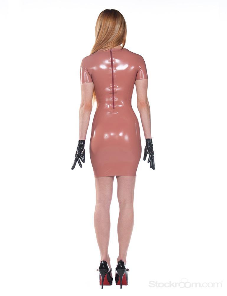 A blonde woman wearing the Jane Latex Dress by Syren Latex in light brown stands turned away from the camera in front of a blank background. She also wears black latex driving gloves and black high heels.