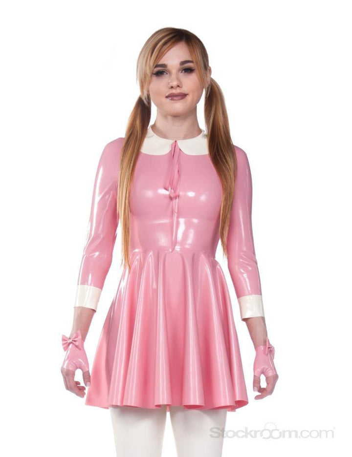 A blonde woman with pigtails wearing the Wednesday Swing Dress by Syren Latex in pink poses in front of a blank background. The dress has three-quarter sleeves, a white peter pan collar with latex ribbons, and white cuffs on the sleeves.