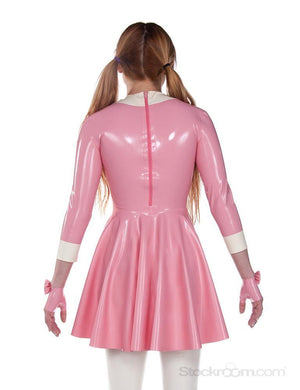 A blonde woman with pigtails wearing the Wednesday Swing Dress by Syren Latex in pink poses in front of a blank background, facing away from the camera. The dress has a zipper up the back.