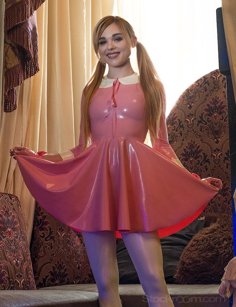 A blonde woman with pigtails wearing the Wednesday Swing Dress by Syren Latex in pink poses in front of an ornate couch. 